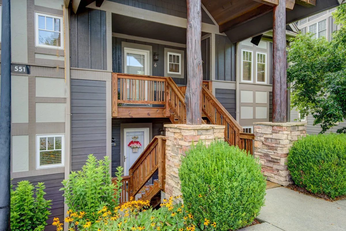 A welcoming exterior view featuring wooden stairs leading up to the condo's entrance, framed by sturdy stone pillars, lush greenery, and vibrant flower beds. The inviting pathway makes a strong first impression and hints at the peaceful mountain surroundings.
