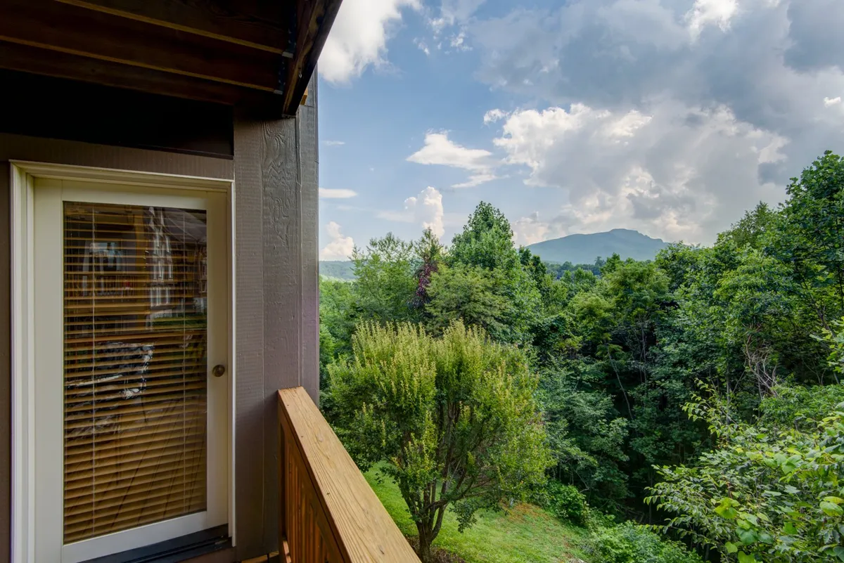 A balcony with a sturdy railing overlooks a vast expanse of green foliage and a distant mountain landscape under a dramatic sky filled with clouds. The scene evokes a peaceful moment, inviting viewers to enjoy the changing seasons and expansive views.