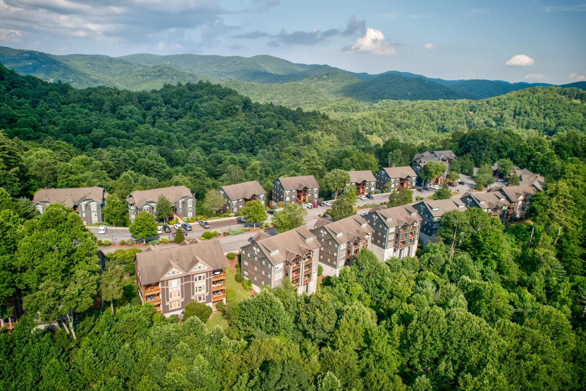 A peaceful scene featuring apartment buildings set amidst lush green trees and a striking mountain backdrop. The landscape feels serene, perfect for both community events and quiet outdoor time, while the mountains hint at winter sports and summer hikes alike.