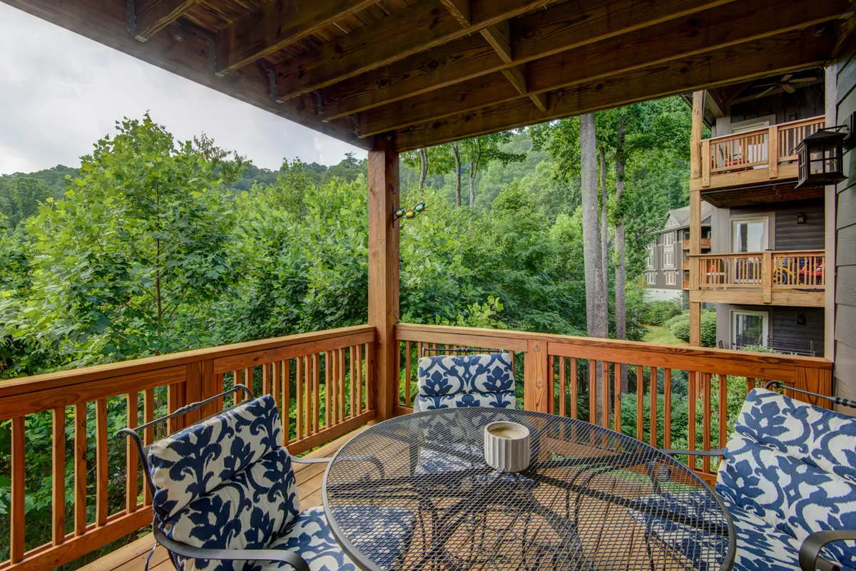 A wooden deck furnished with a circular table and blue-and-white chairs is nestled amidst lush greenery. The serene arrangement offers a cozy spot for reading, dining, or simply enjoying the surrounding nature, with a view into the treetops and hints of the mountainous landscape beyond.