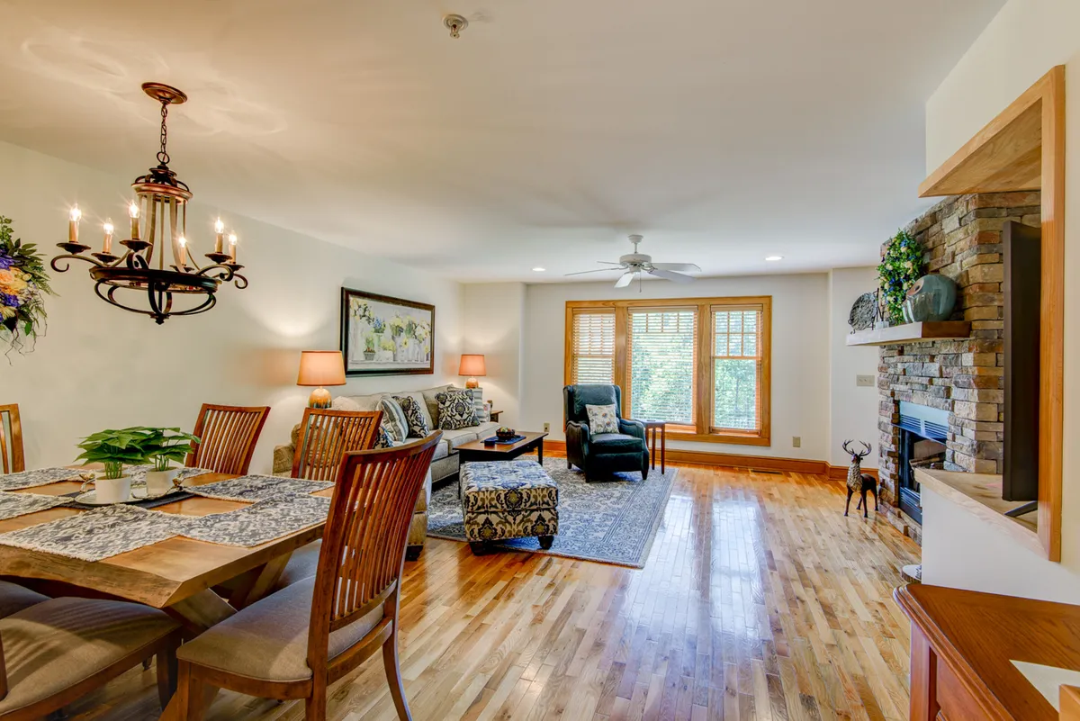 A spacious, open-plan living and dining area featuring a large, comfortable sofa, a striking fireplace, a wooden dining table under a chandelier, and expansive windows that fill the space with natural light. The scene evokes a welcoming, communal atmosphere perfect for gatherings.