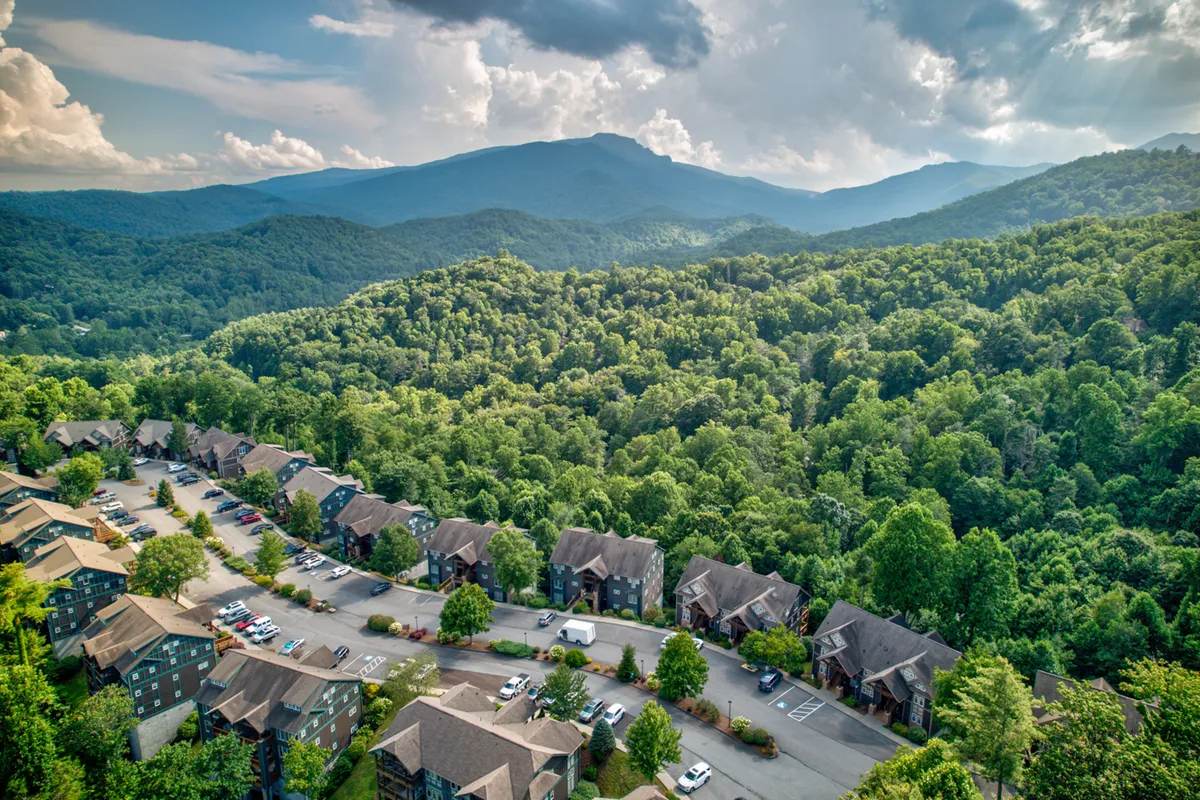 A sweeping view of the mountains surrounded by dense forests, with residential buildings nestled among the trees and a curved road winding through the landscape. The scene is tranquil, highlighting the natural beauty and outdoor recreational opportunities in the area.