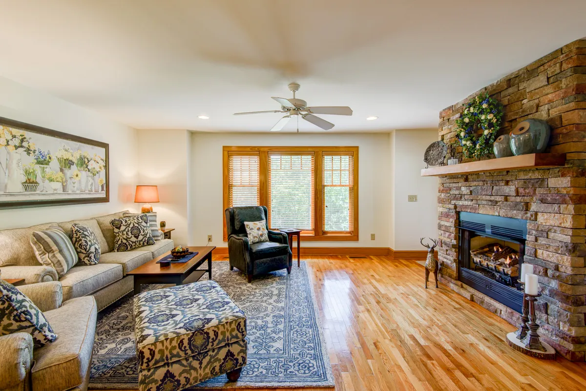 A close-up view of a comfortable sofa adorned with decorative pillows, positioned beside a striking stone fireplace and large windows, with sunlight streaming in. The ambiance is both cozy and bright, emphasizing relaxation.