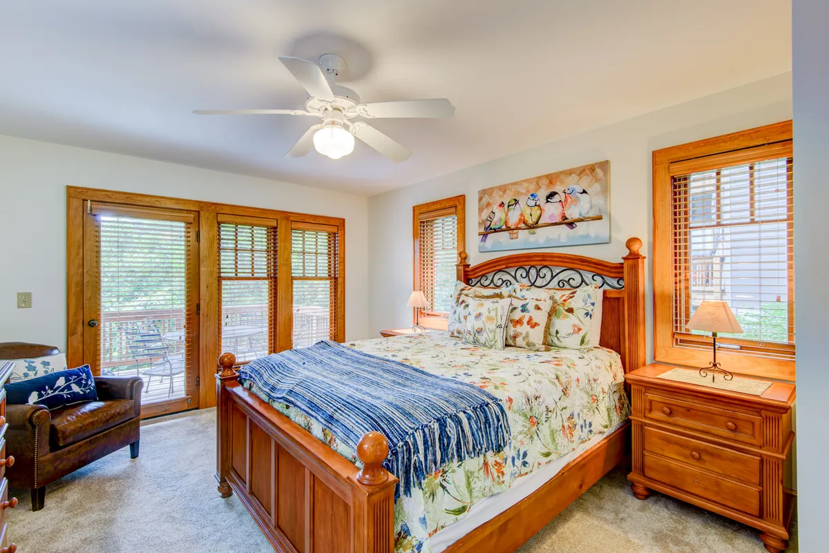 This image shows a serene bedroom featuring a bed with decorative pillows, wooden furniture, and open balcony doors letting in natural light. The tranquil mood is underscored by soft lighting and a peaceful color palette, creating an inviting retreat ideal for relaxation.
