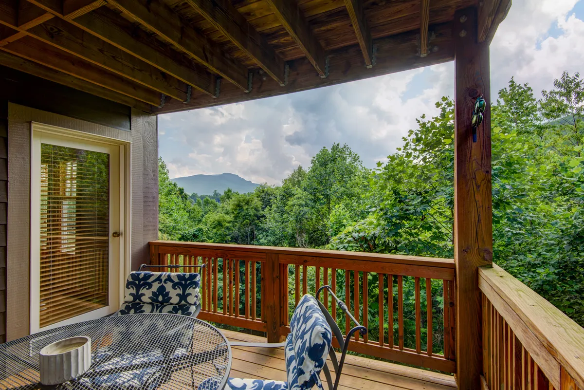 A wooden deck with decorative chairs and a table offers an inviting setting to relax and socialize. The deck overlooks a sweeping mountain vista, with layers of peaks visible in the distance and lush greenery below. The setup suggests both sunrise and sunset views, with ample outdoor seating for gatherings or quiet moments.