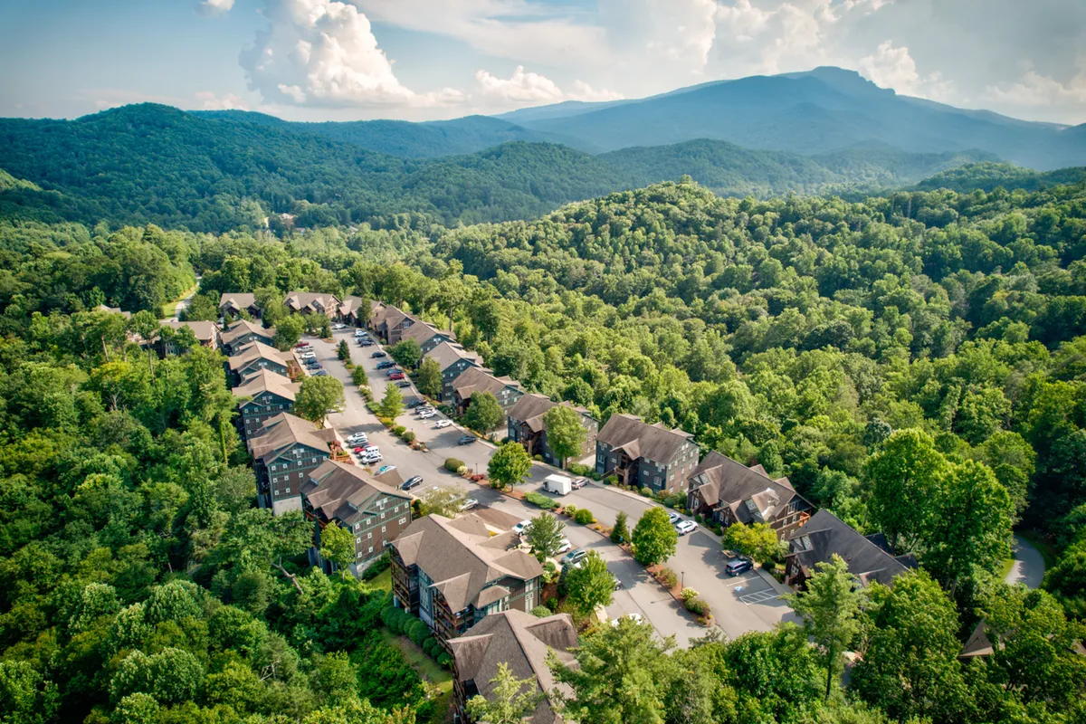 Rows of residential buildings situated among lush green trees, with mountains in the distance and neatly arranged parking spaces in the foreground. The organized, peaceful atmosphere evokes a sense of retreat and community activity.